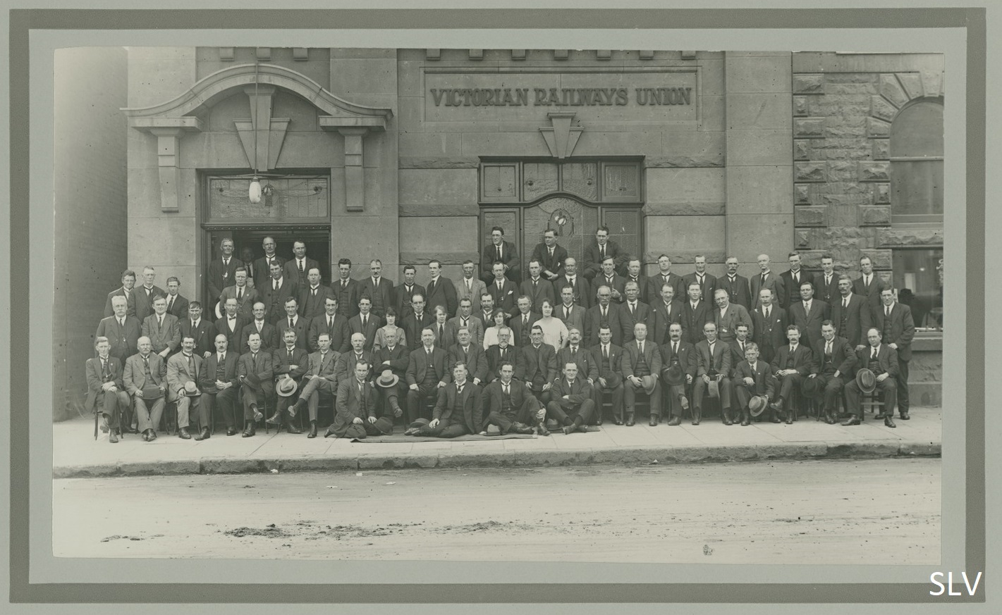 Group of men with two women standing in centre of third row, same building in background. Wall behind them with sign above window reading Victorian Railways Union