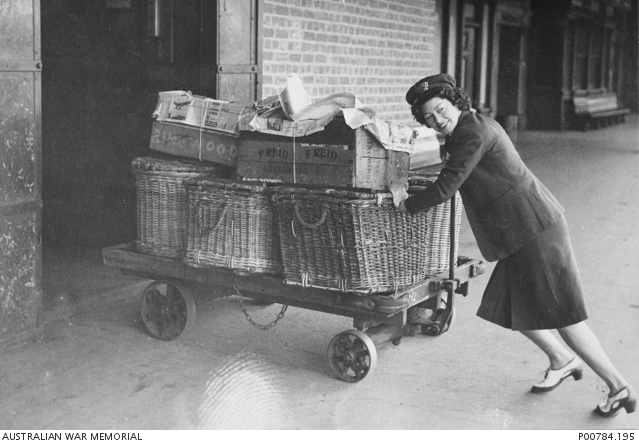 A woman railway porter with the Victorian Railways pushes a trolley laden with wicker baskets and boxes into a building at a railway station.