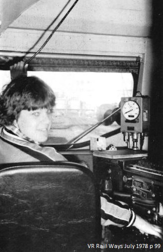 A woman in locomotive driver’s cab. Woman is sitting in driver’s seat and turn back to look at the camera. There are driver’s controls to her right, she has hand on the horn cable over her head.