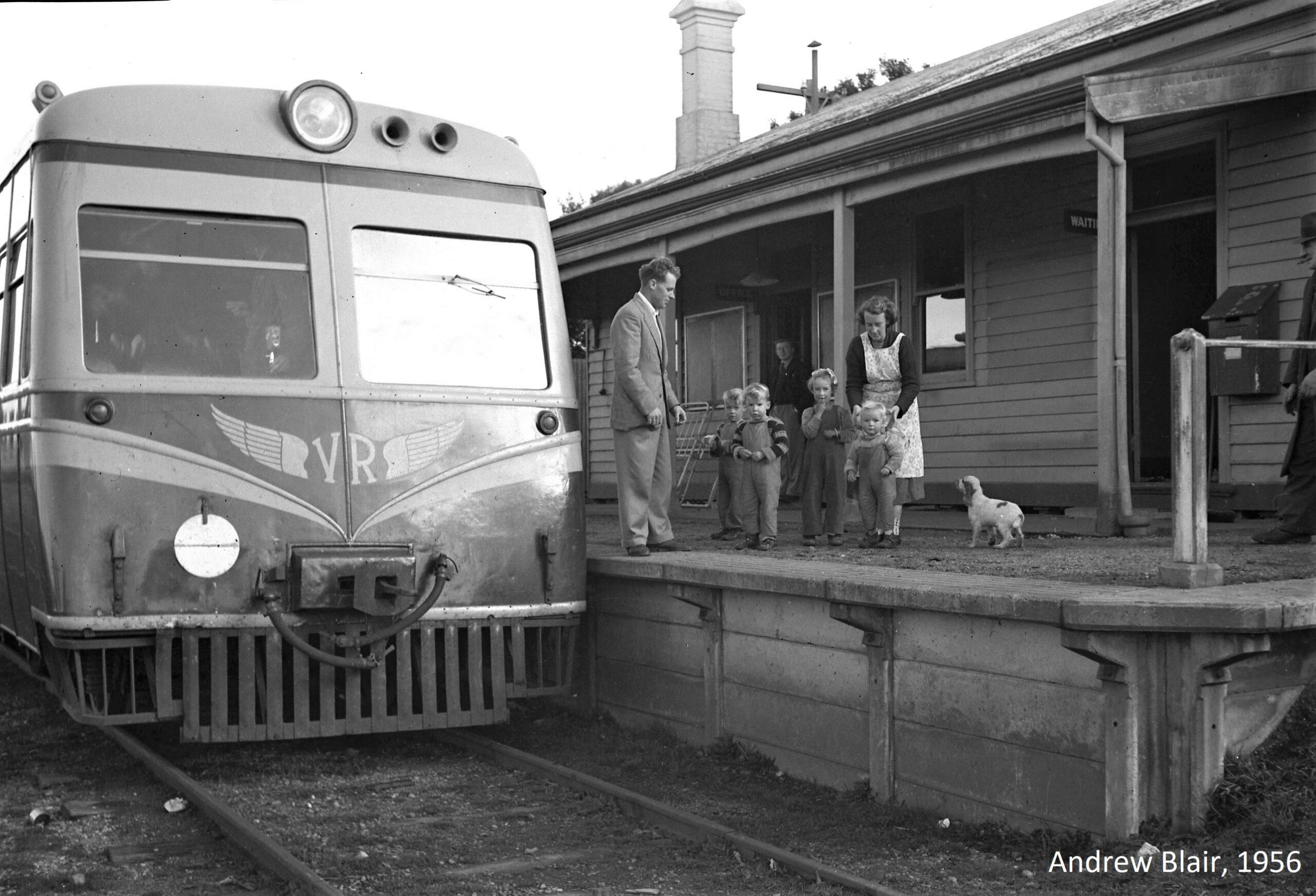 Woman wearing an apron standing on railway station platform with four small children in front of her and a dog to her side. A man in a suit is approaching her. A Victorian Railways rail motor train is stationary at the platform.