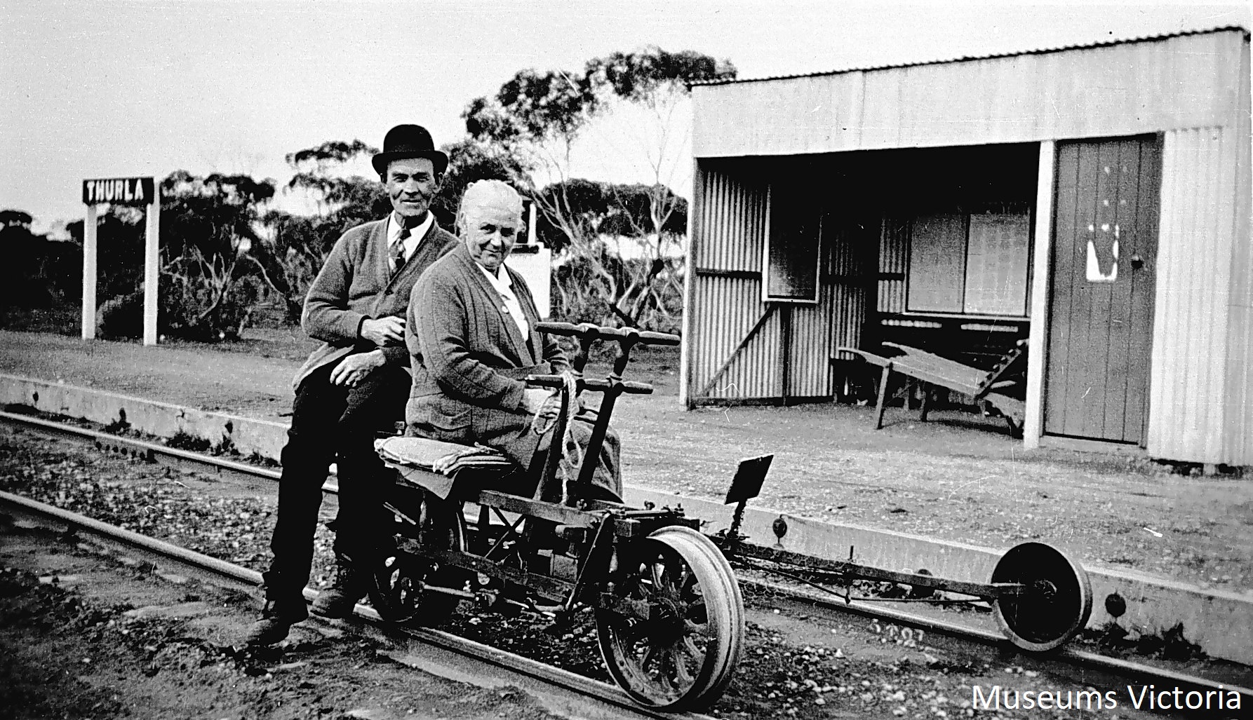 The stationmistress and ganger of Thurla Railway Station posed on a ganger's trolley. They were husband and wife. A small corrugated iron shelter and the station sign are visible behind them. A wooden construction, possibly a handcart of some kind, in the shelter.