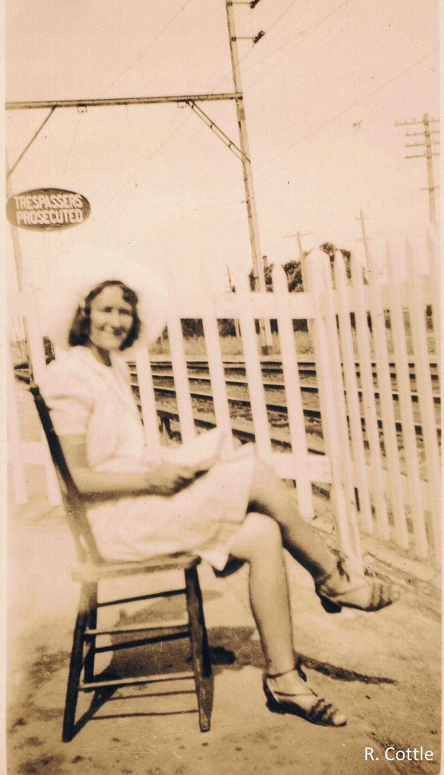 Woman wearing dress and sandals seated at railway gates waiting for trains. Railway tracks in background. Sign reads ‘TRESPASSERS PROSECUTED’.