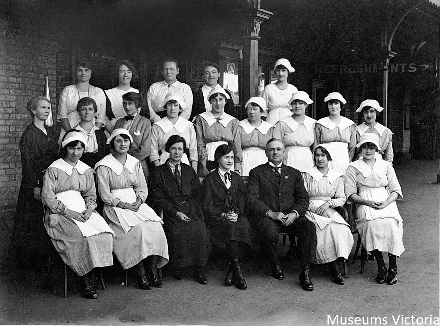 A group of 21 refreshment room staff pose on the platform at Bendigo Railway Station where Mondays were a very busy morning with passenger trains arriving from Echuca, Swan Hill, Heathcote, Cohuna, Robinvale & Kulwin. The sole male staff member seated in the front row is the Manager, while the cook appears (in black) at the left-hand end of the middle row.