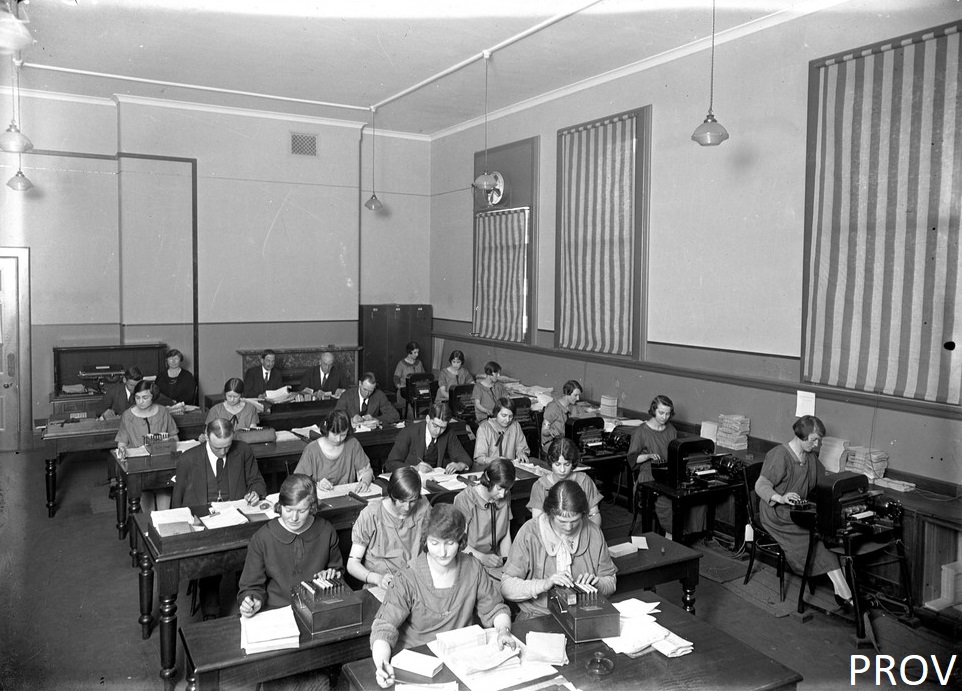 Office with rows of desks with women and a few men seated at accounting machines, working with their heads down. Large windows covered with drapes with vertical stripes.